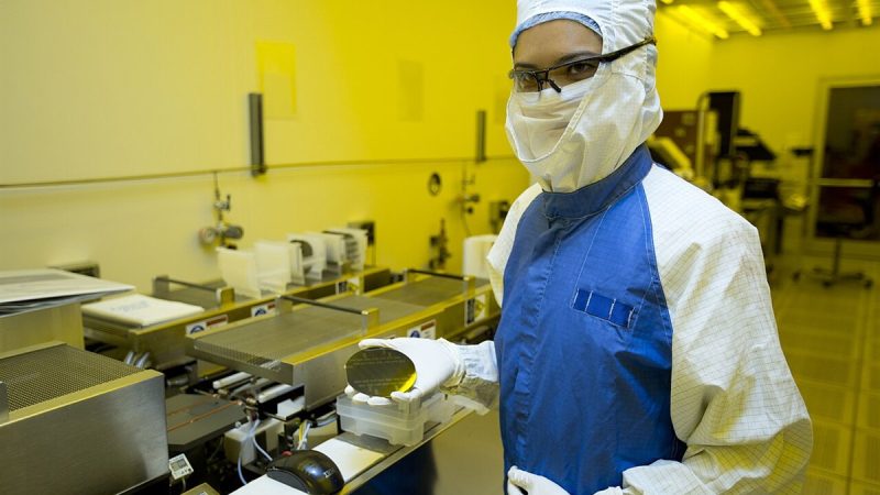 A technician in cleanroom clothing holding a silicon wafer in a semiconductor fabrication plant